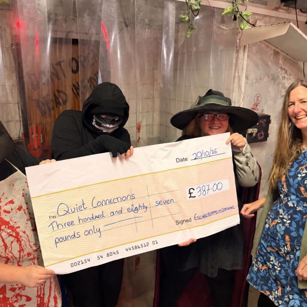 A giant cheque for £387 made out to Quiet Connections, held by three costumed escape room characters and a representative of Quiet Connections inside an escape room set.